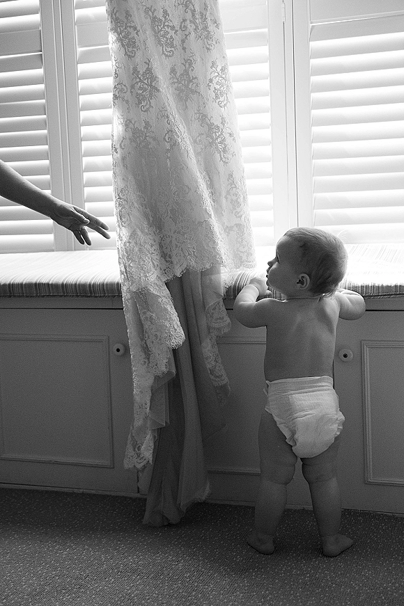 Wedding dress detail, lace wedding dress hanging on a hanger by window shutters as a baby sits on the bench reaching toward the hem