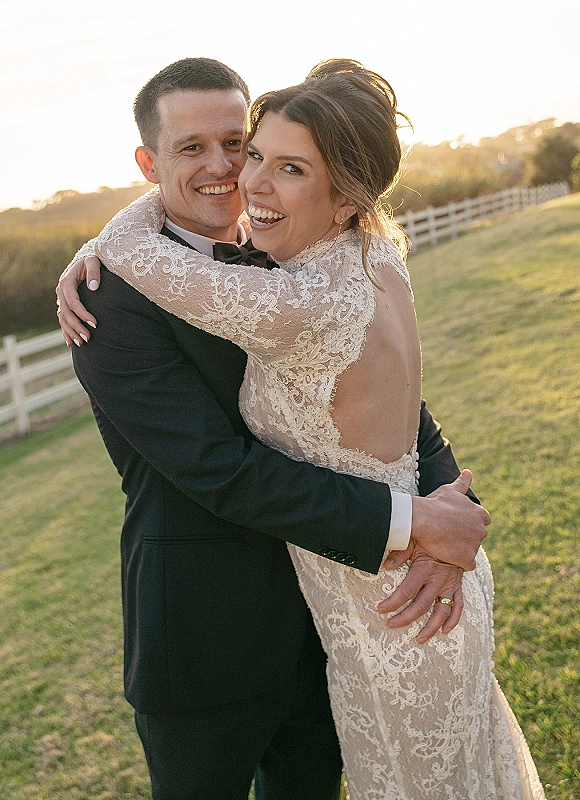 Couple portrait of bride hugging groom, laughing in a grassy field by a wooden fence at sunset, with her open-back lace gown visible