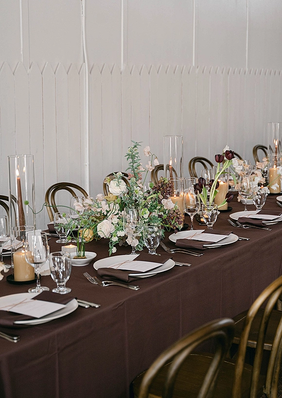 Reception tablescape with long banquet table decor featuring dark linens, low roses and tulips, and candlelight against a white paneled wall