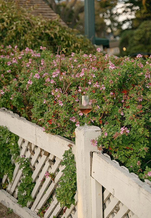 Wine glass detail with rosé wine glass and pink flowers resting on a white wooden fence, set against lush garden greenery and blooms