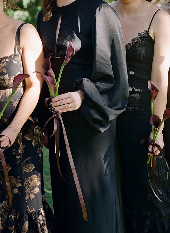 Bridesmaid dresses in black satin and lace, with sheer sleeves, holding dark calla lily bouquets tied with ribbon in sunlit greenery