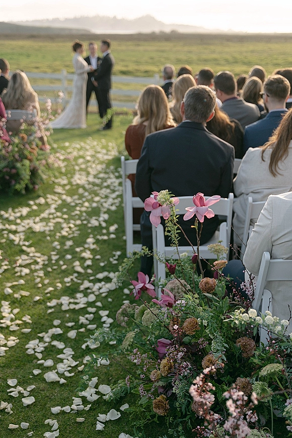 Outdoor wedding ceremony with white folding chairs and a flower petal aisle on grass, framed by pink and white roses by open hills