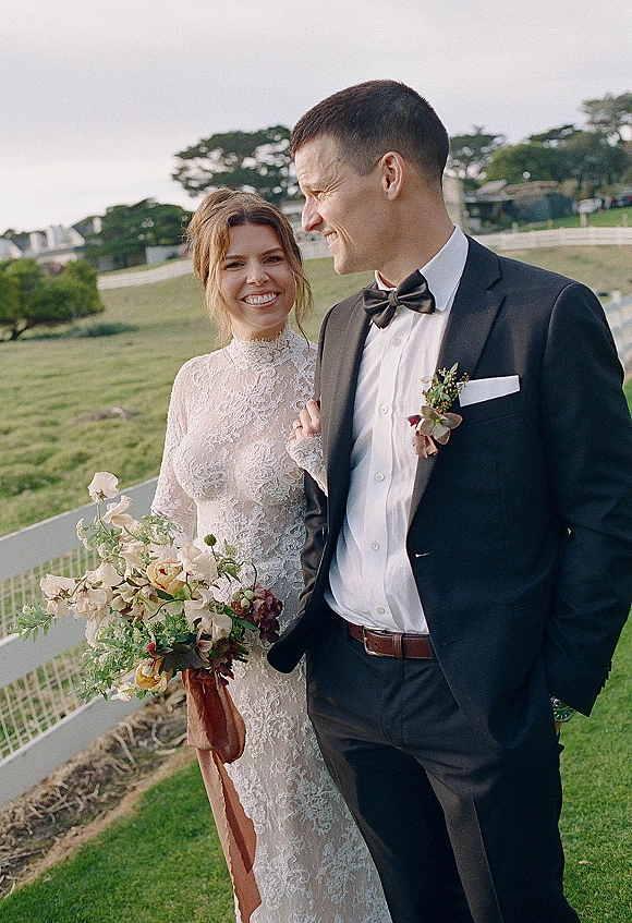 Couple portrait of bride and groom walking arm in arm on a grassy lawn, bride holding bouquet, with white fence and trees behind