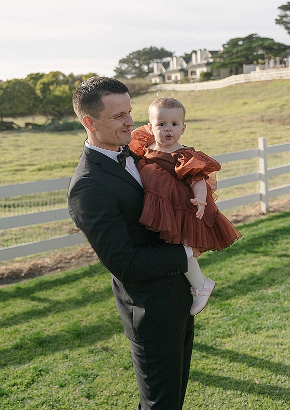 Groom with child in a black tuxedo and bow tie holding a toddler in a tulle-sleeve dress on a grassy lawn by a white fence