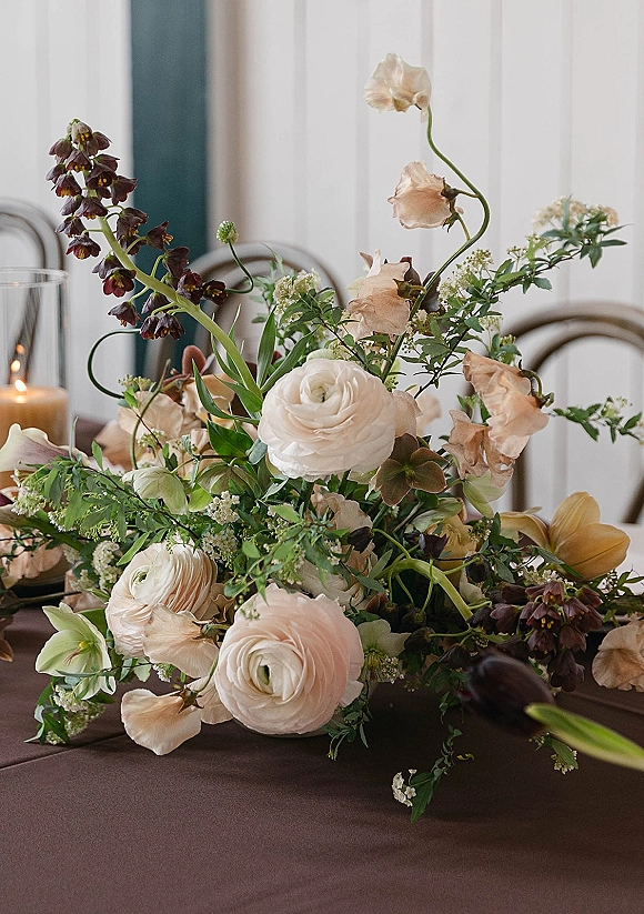 Wedding centerpiece with ranunculus and sweet peas in a compote, greenery and hurricane candle on linen, softly lit by window light