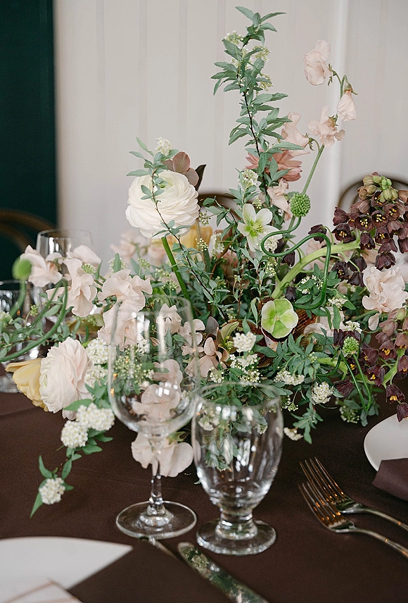 Wedding centerpiece with garden style centerpiece blooms in blush, white, and burgundy, set among greenery, plates, silverware, and glasses.