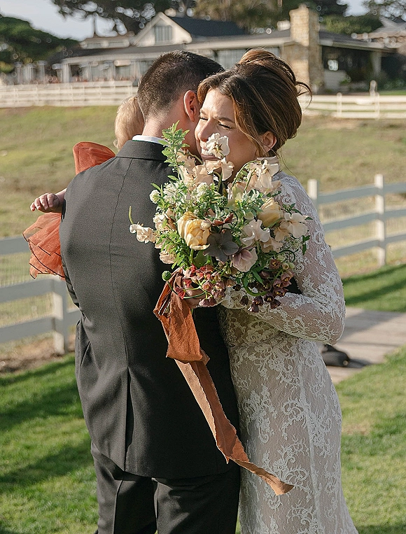 Wedding hug as bride hugs groom, holding a rose bouquet with silk ribbon in front of a white fence and farmhouse lawn setting