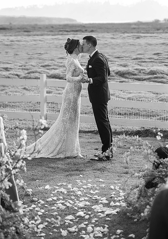 Wedding kiss as bride in lace long-sleeve dress and groom in tuxedo hold hands on rose petals by a white fence and open field