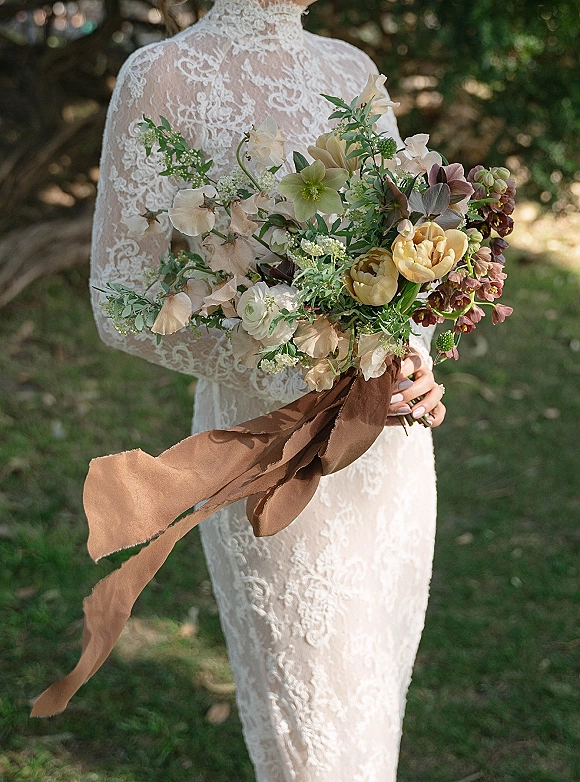 Bridal bouquet with garden bridal bouquet blooms and greenery, held by a bride in a lace long sleeve dress on a grassy garden lawn with trees