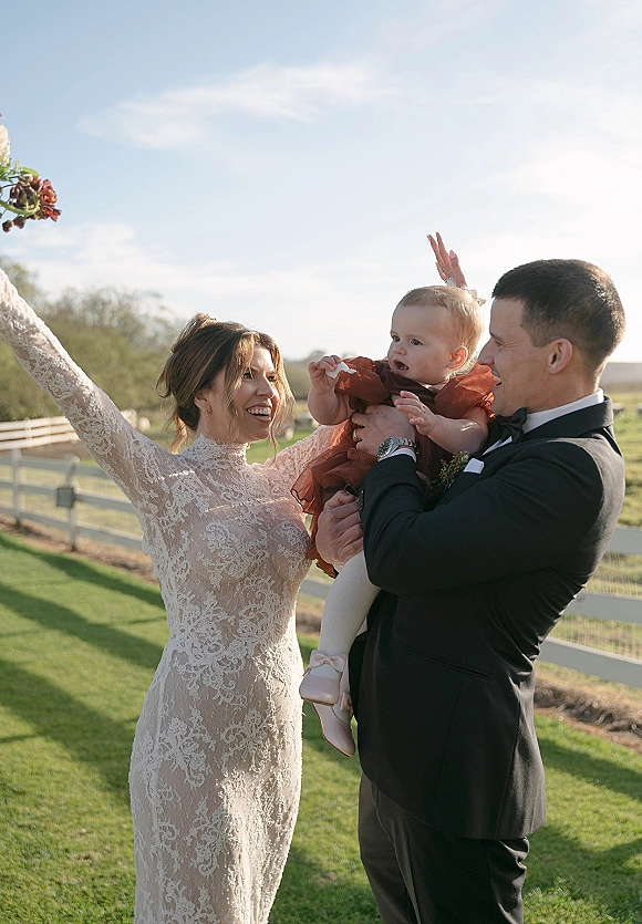 Family wedding portrait of bride and groom with baby, bride in long sleeve lace dress, on a lawn by a white fence under blue sky