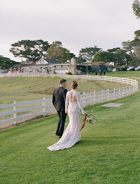 Couple portrait of bride and groom holding hands, walking away on a green lawn toward a white fence, lace open-back gown and bouquet ribbon