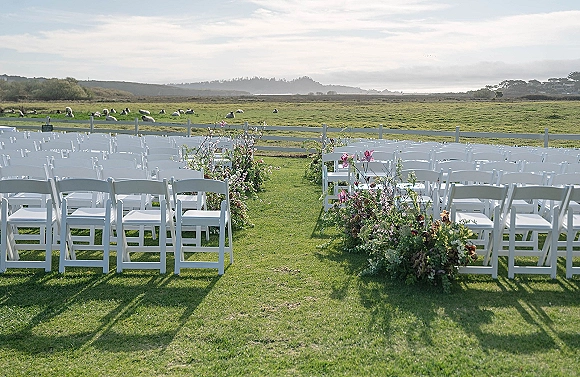 Ceremony seating with white folding chairs and aisle florals on a grass lawn, set before a white fence with grazing sheep and hills
