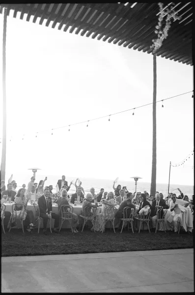 Reception moment at an outdoor wedding reception beneath string lights, with the ocean and palm trees framing the lawn.