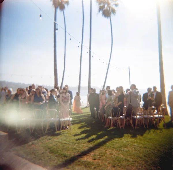 Ceremony moment at an outdoor wedding ceremony as guests line a simple aisle under string lights, with palm trees and ocean beyond.