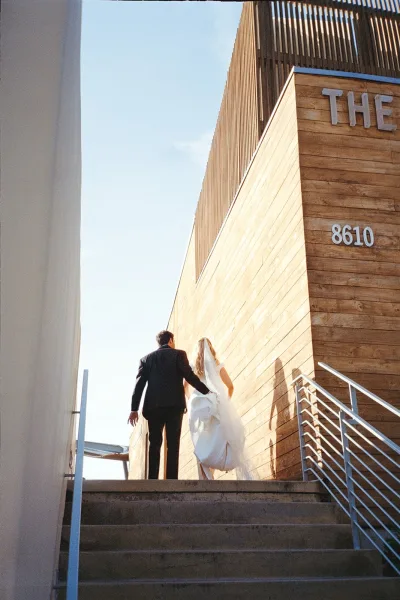 Couple portrait of a bride and groom walking up concrete steps, her veil trailing, against a wood wall and metal railing under a blue sky.