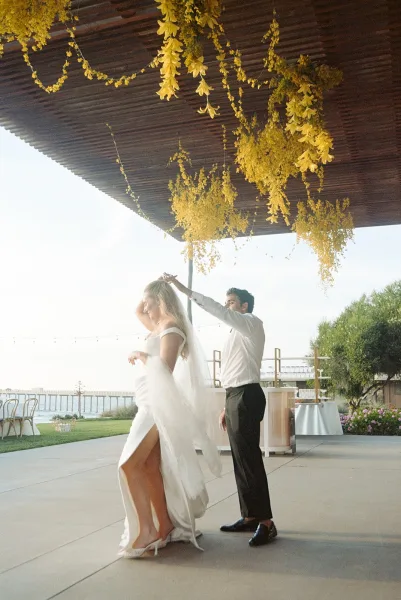 Couple dancing their first dance on a waterfront patio under string lights, framed by a wooden canopy and trees swaying beside the pier.
