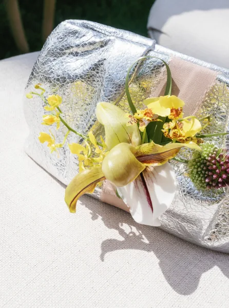 Bridal clutch, a silver wedding clutch adorned with orchids, rests on linen fabric against a grass lawn backdrop.