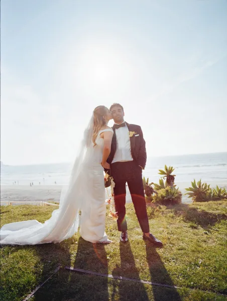Couple portrait, a wedding kiss portrait on a sunlit beach lawn with ocean beyond, the bride’s veil floating as they embrace.