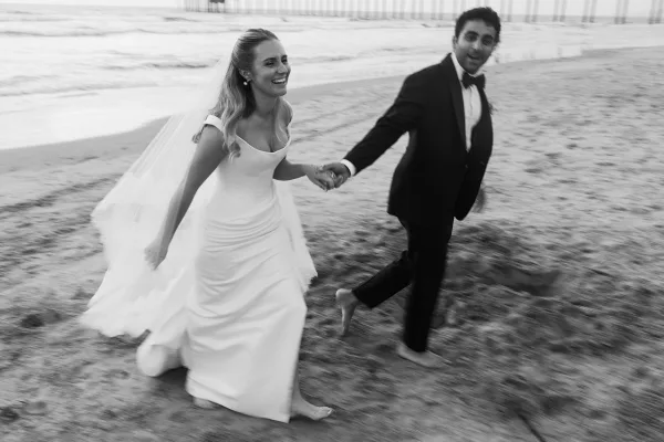 Couple portrait of the bride and groom running hand in hand along a sandy shoreline, her veil streaming as ocean waves roll by near a wooden pier.