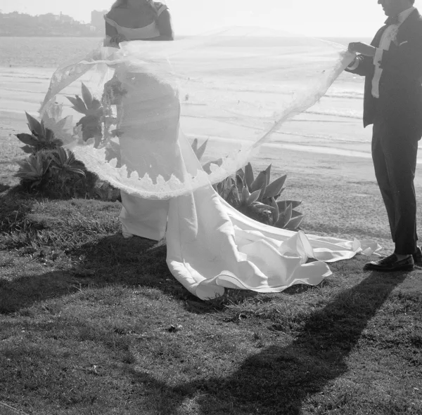 Bridal portrait in black and white captures a bride with a flowing veil on a sandy beach with the ocean behind her.