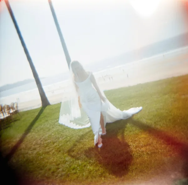 Bridal portrait of a beach bride with a long train on a sunlit lawn by the ocean, framed by swaying palm trees and chairs.