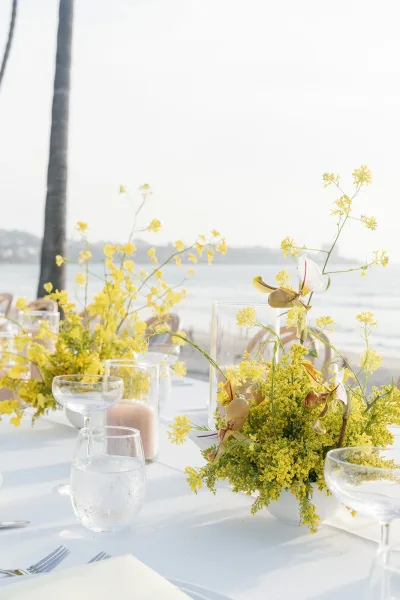 Reception tablescape in beach reception decor with airy floral centerpieces, set by the ocean with a palm tree, distant shoreline, and open sky.