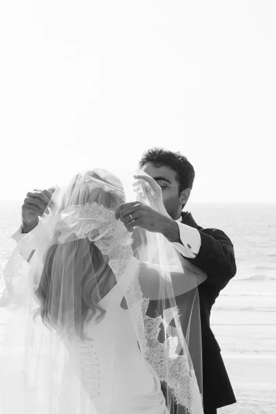 Veil lift moment as an under the veil shot frames the couple beneath a lace veil, sharing an embrace on a breezy beach by the ocean.