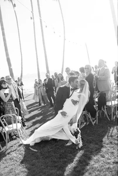 Ceremony exit with a ceremony recessional kiss as the groom dips the bride, her veil trailing, guests cheering beneath palm trees by the ocean.