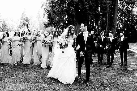 Wedding party portrait of the bride and groom walking with bridesmaids and groomsmen on a grassy lawn with tall trees, bride holding bouquet