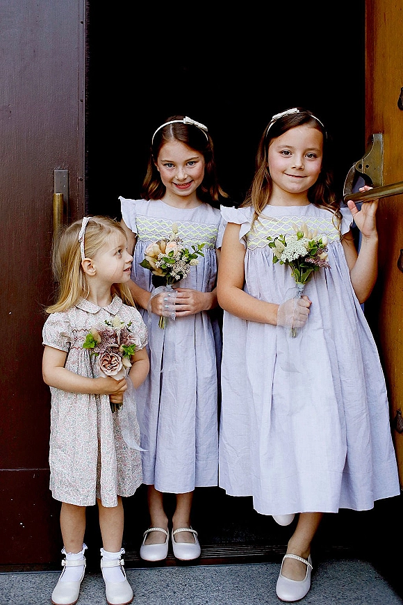 Flower girls in white dresses holding bouquets with hair bows, standing together in a wooden barn doorway during a rustic wedding