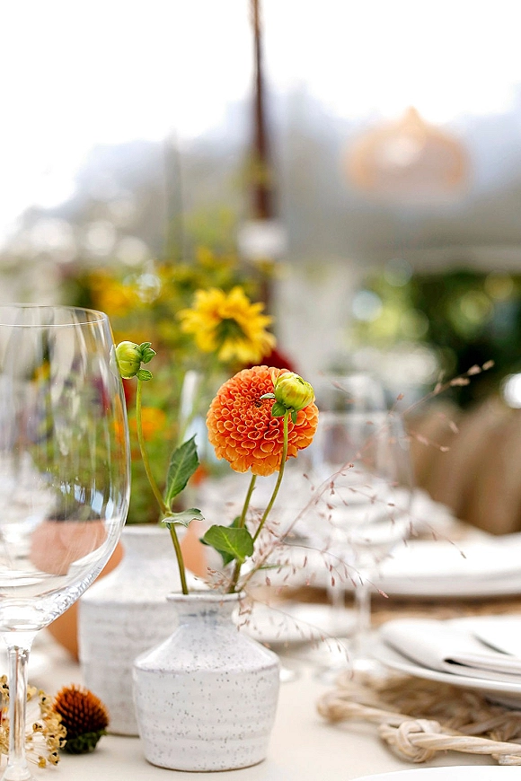 Reception tablescape with an orange dahlia bud vase centerpiece, taper candles and woven charger place settings on an outdoor patio under umbrellas