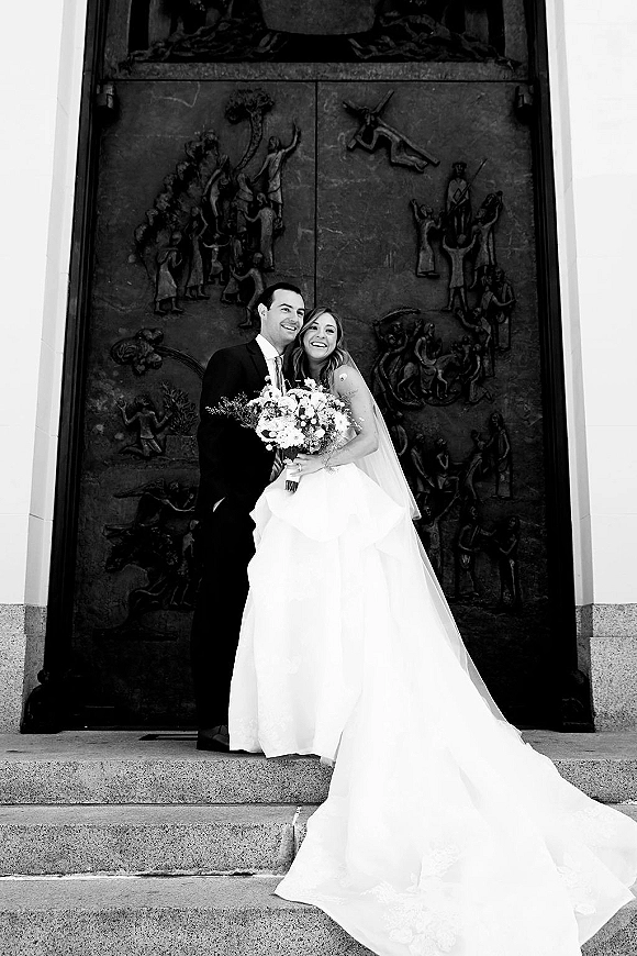 Couple portrait of bride and groom laughing, her veil and white bouquet visible, posed on stone steps before an ornate carved door