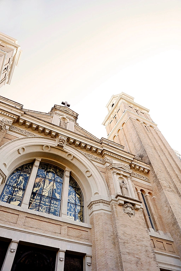 Church exterior with a stained glass window and arched entryway beneath a bell tower and cross, photographed from below against the sky