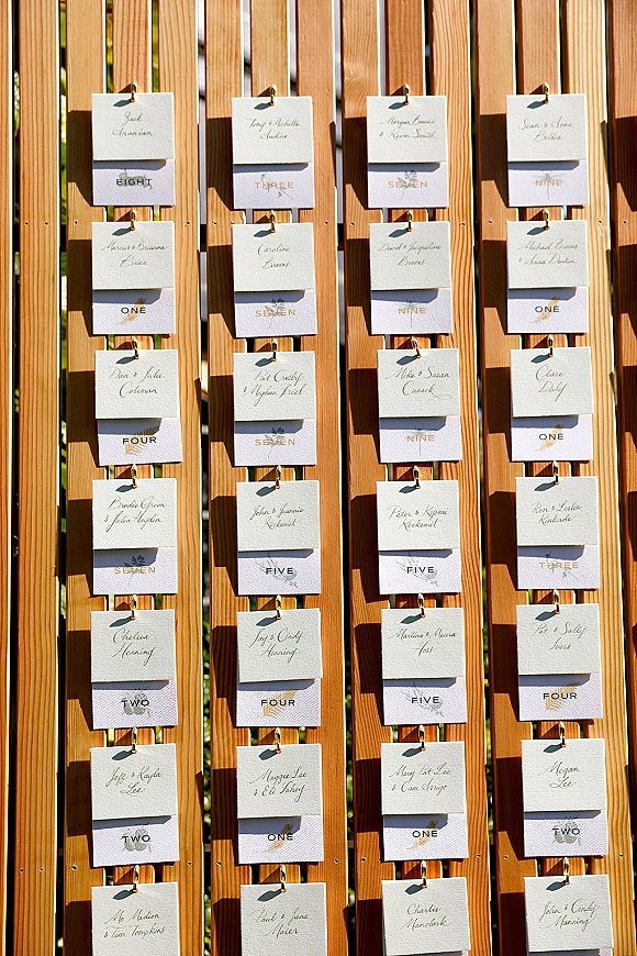 Wedding escort cards clipped with gold clips to a wooden slat display, featuring calligraphy and table numbers in sunlight outdoors