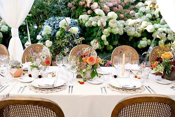 Reception tablescape with an outdoor wedding table set in white linens, woven placemats, colorful centerpieces, and glass hurricane candlelight in a garden with hydrangeas and white drapery