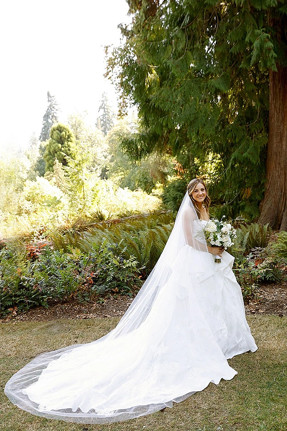 Bridal portrait of a bride holding bouquet in a strapless gown with a long veil, standing in sunlit garden greenery with ferns