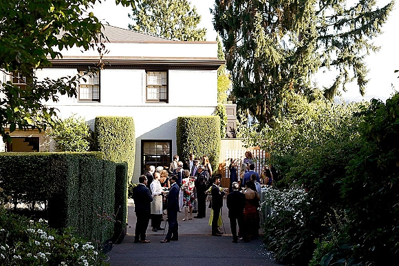 Wedding cocktail hour with guests mingling outdoors, holding cocktail glasses in suits and dresses along a hedge-lined garden pathway