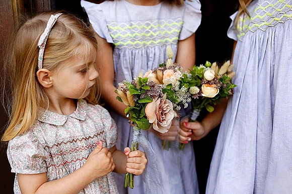Flower girl holding a flower girl bouquet of pastel blooms and greenery, wearing a smocked dress and headband against a dark indoor backdrop