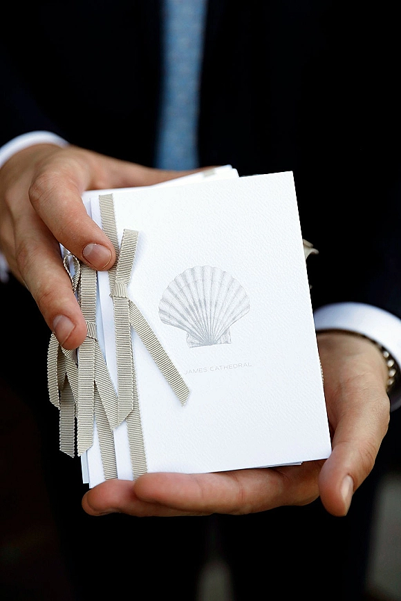 Wedding invitation with minimalist wedding invitation design, tied with a ribbon bow and shell motif on textured paper against dark suit fabric