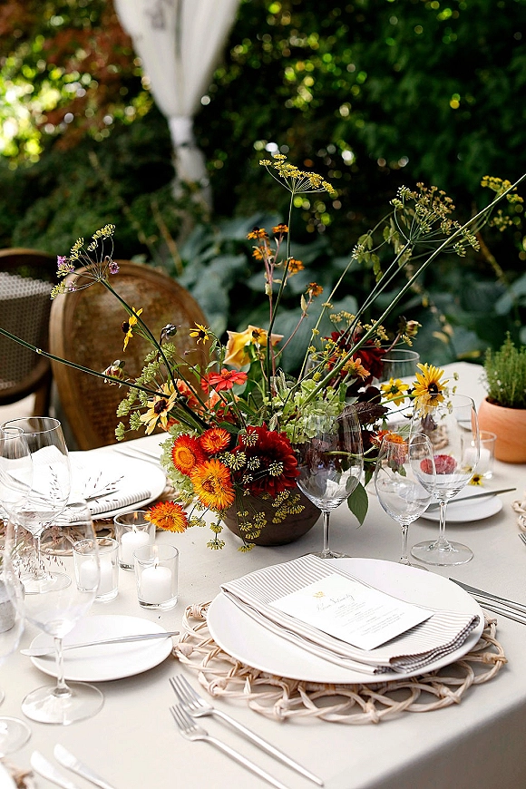 Reception tablescape with wildflower wedding centerpiece in a ceramic bowl, taper candles, striped napkins, and menus on plates on a garden patio