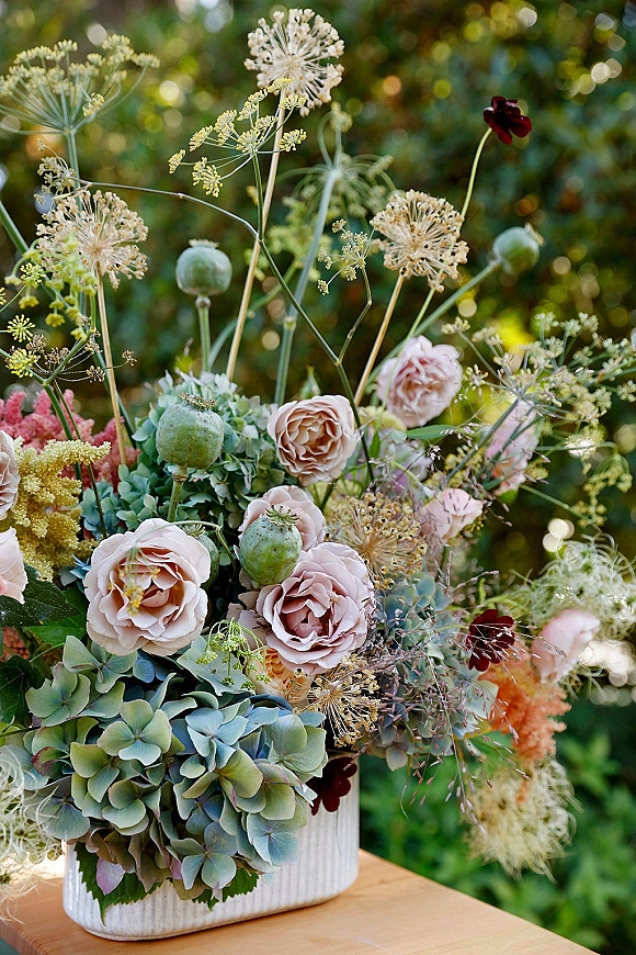 Wedding centerpiece flowers in a garden wedding centerpiece, featuring blush roses, blue hydrangea, greenery, and poppy pods in a ceramic vase.