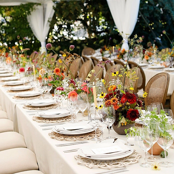 Reception tablescape with wildflower wedding centerpieces, taper candles, woven chargers, and place cards under a draped garden tent canopy