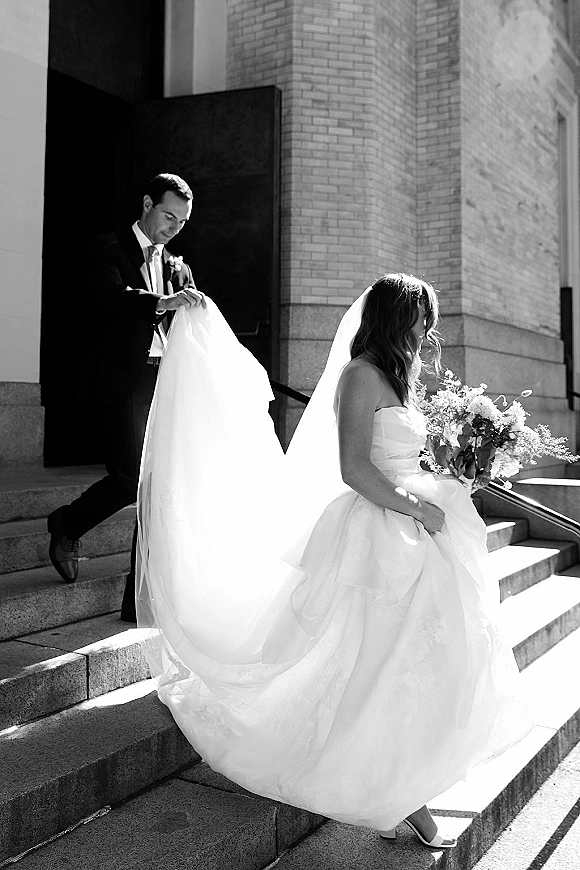 Wedding couple portrait of bride walking up stone steps with bouquet and veil, groom holding her long dress train by a sunlit brick doorway