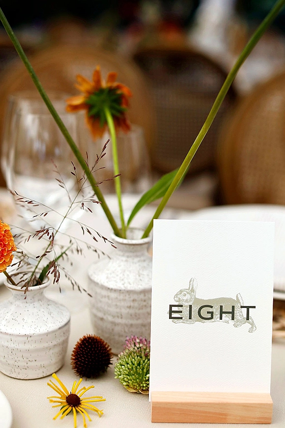 Wedding table number and table number card in a wooden holder beside ceramic bud vases with orange flowers and dried grasses on white linen