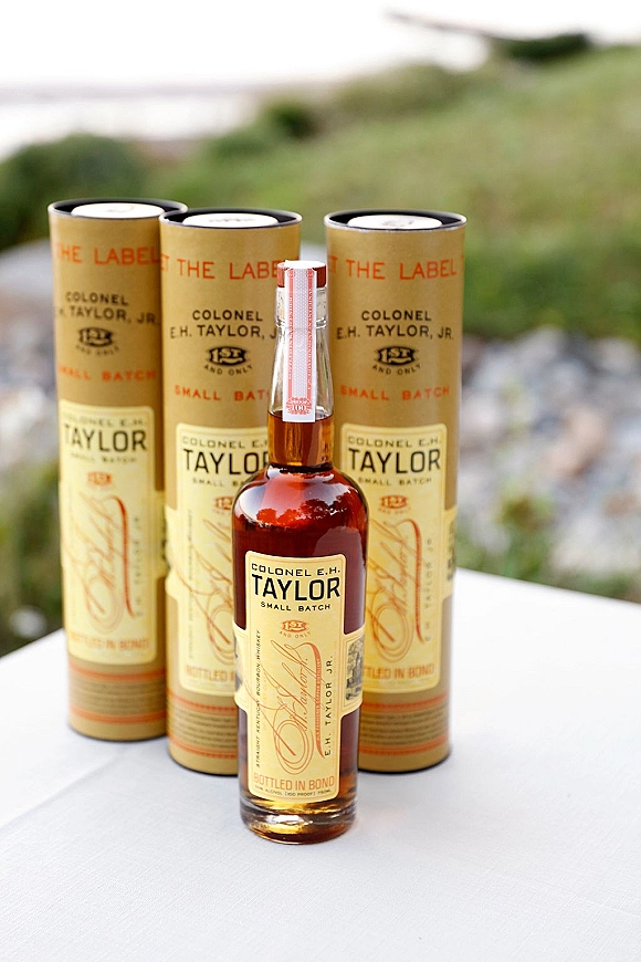 Wedding whiskey favors arranged with mini whiskey bottle favors in clear tubes on a tablecloth beside an outdoor lawn and water backdrop