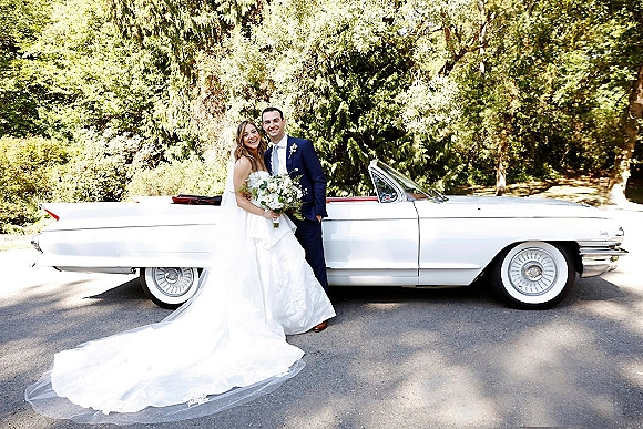 Couple portrait of bride and groom by car, smiling beside a classic convertible on a sunlit tree-lined road, bride holding white bouquet