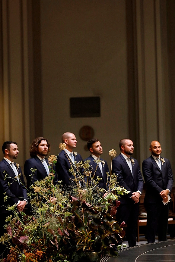 Groomsmen lineup in navy suits with boutonnieres and ties standing at the altar before draped curtains and wooden benches indoors