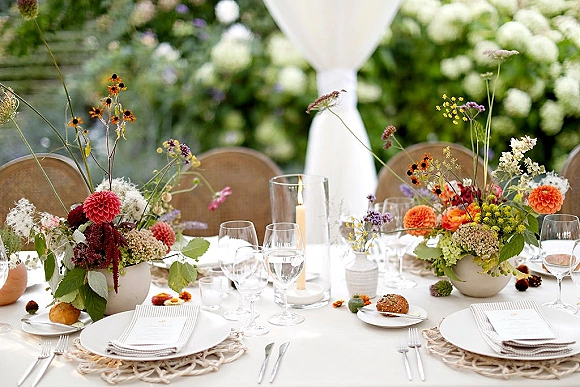 Reception tablescape with wedding table centerpiece of wildflower blooms, pillar candles, and woven placemats against white drapery and greenery backdrop