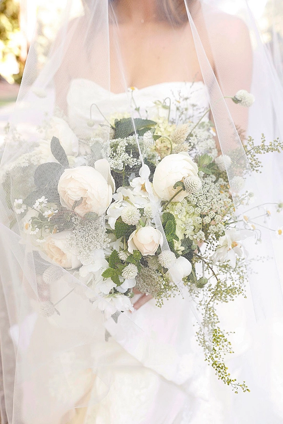 Bridal bouquet of white roses and orchids with baby's breath and greenery beneath a sheer veil in soft sunlit outdoor greenery
