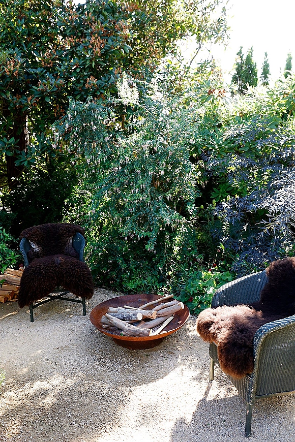 Outdoor lounge area with wedding lounge seating, wicker chairs draped in sheepskin around a fire pit bowl on a gravel patio amid garden trees